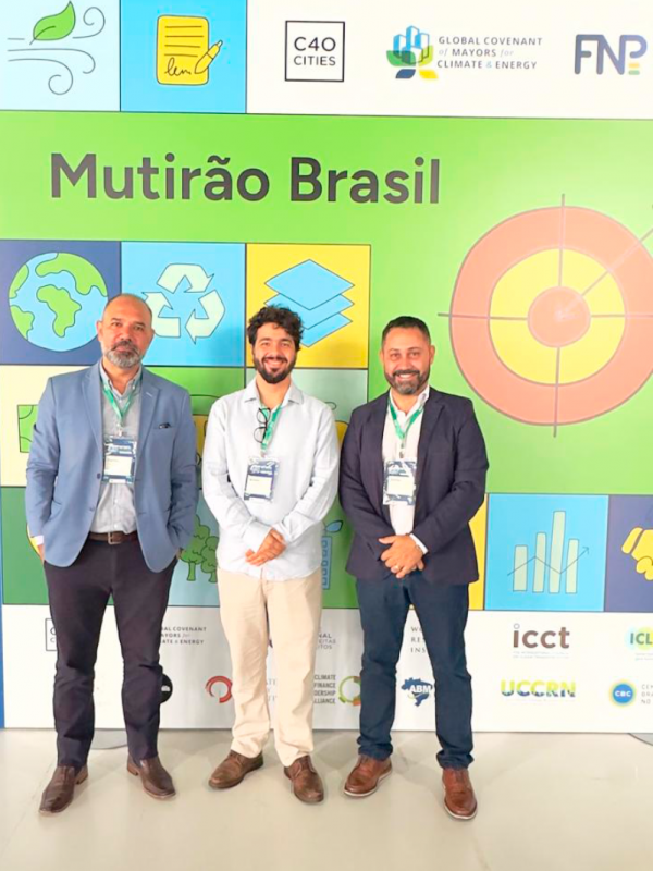 Tr&ecirc;s homens adultos est&atilde;o em p&eacute;, lado a lado, posando para uma foto em um ambiente interno. Eles vestem roupas sociais &mdash; blazers, camisas e cal&ccedil;as &mdash; e usam crach&aacute;s pendurados no pesco&ccedil;o. Todos est&atilde;o olhando para a c&acirc;mera, com express&otilde;es neutras a levemente sorridentes.

Ao fundo, h&aacute; um painel colorido com o t&iacute;tulo &ldquo;Mutir&atilde;o Brasil&rdquo; em destaque. O painel &eacute; composto por blocos com ilustra&ccedil;&otilde;es que remetem &agrave; sustentabilidade, como planeta Terra, s&iacute;mbolo de reciclagem, folhas e gr&aacute;ficos. Na parte superior, aparecem logotipos de organiza&ccedil;&otilde;es como C40 Cities, Global Covenant of Mayors for Climate & Energy e FNP.

O ch&atilde;o &eacute; claro e o ambiente &eacute; bem iluminado, com aspecto de evento institucional ou confer&ecirc;ncia.